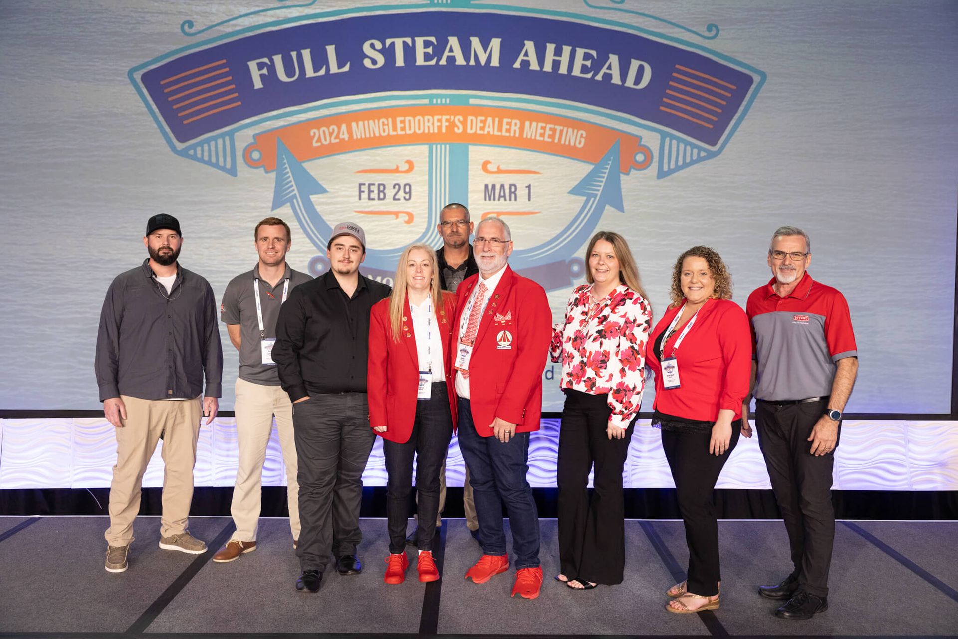 Group of eight people standing onstage in front of a “Full Steam Ahead” event backdrop, some wearing red jackets.