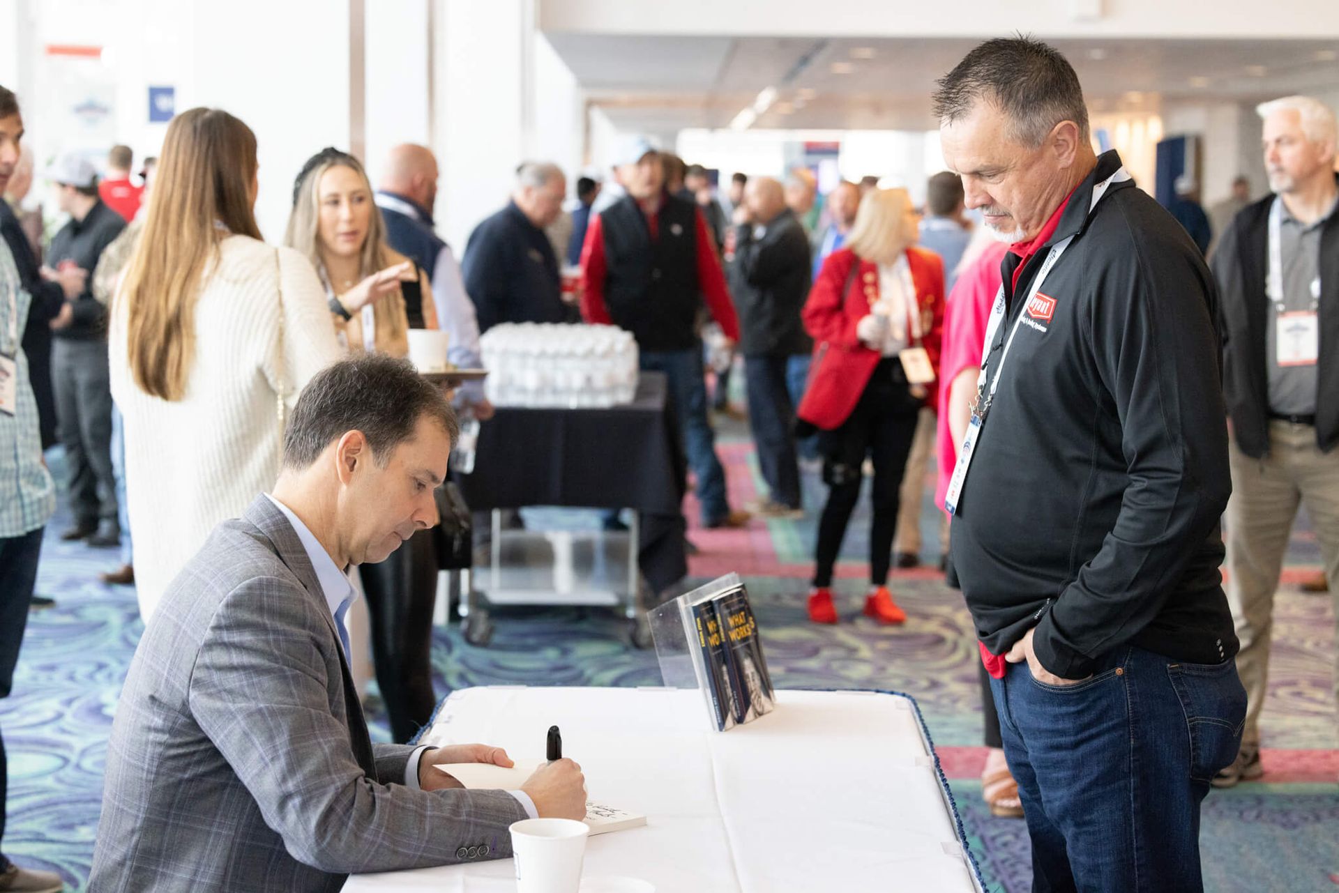 Man signing a sheet at a registration table while another man stands nearby in a busy conference lobby
