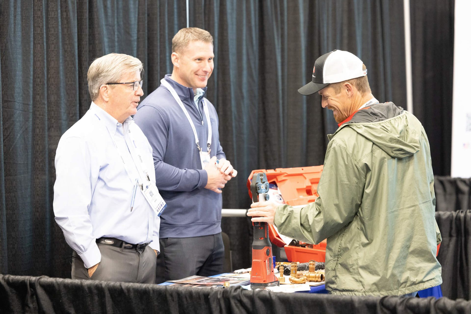 Two men speaking with a worker at a table displaying tools and equipment in an indoor booth