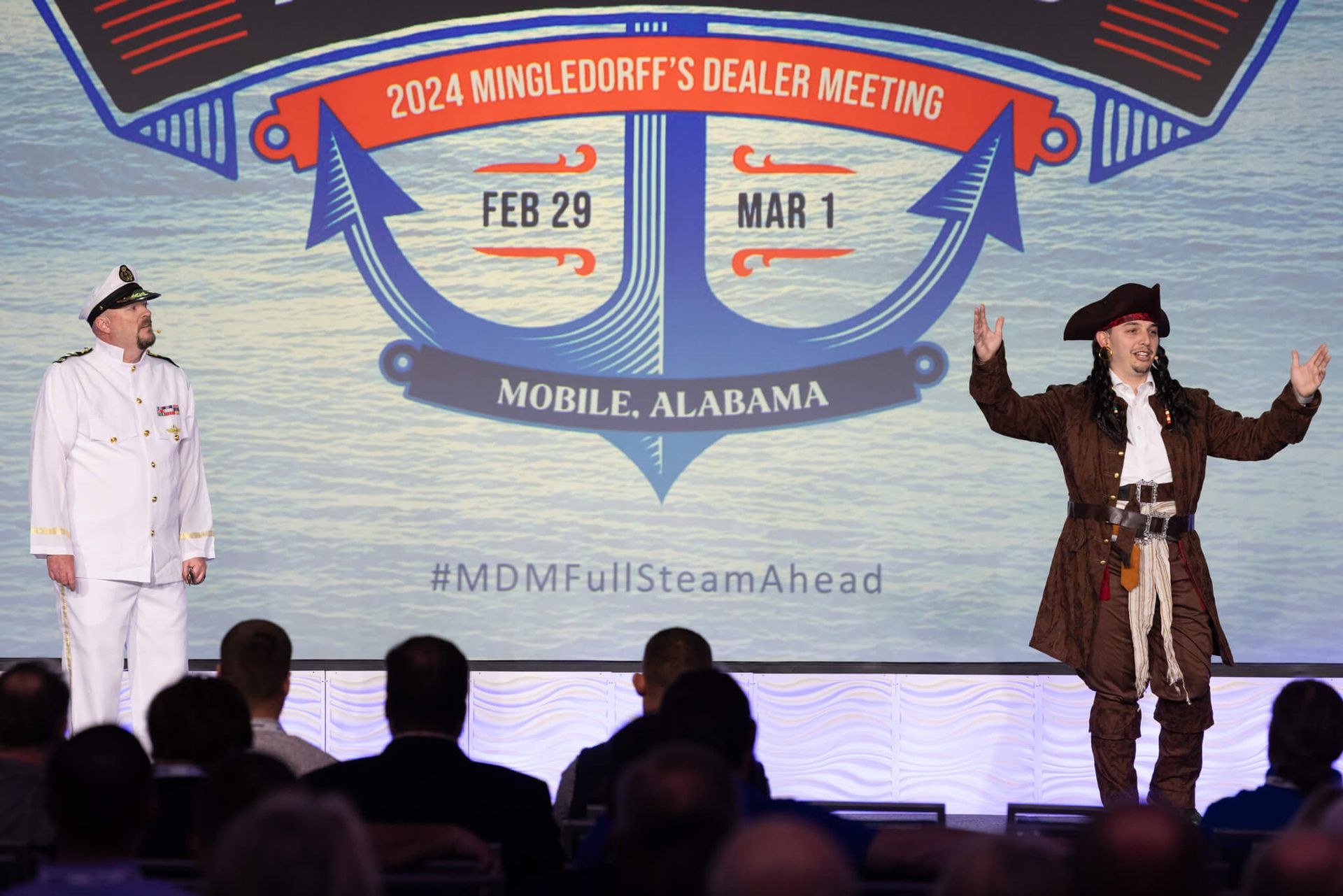 Two costumed performers on a stage beside a MingleDorf's Dealer Meeting banner in Mobile, Alabama