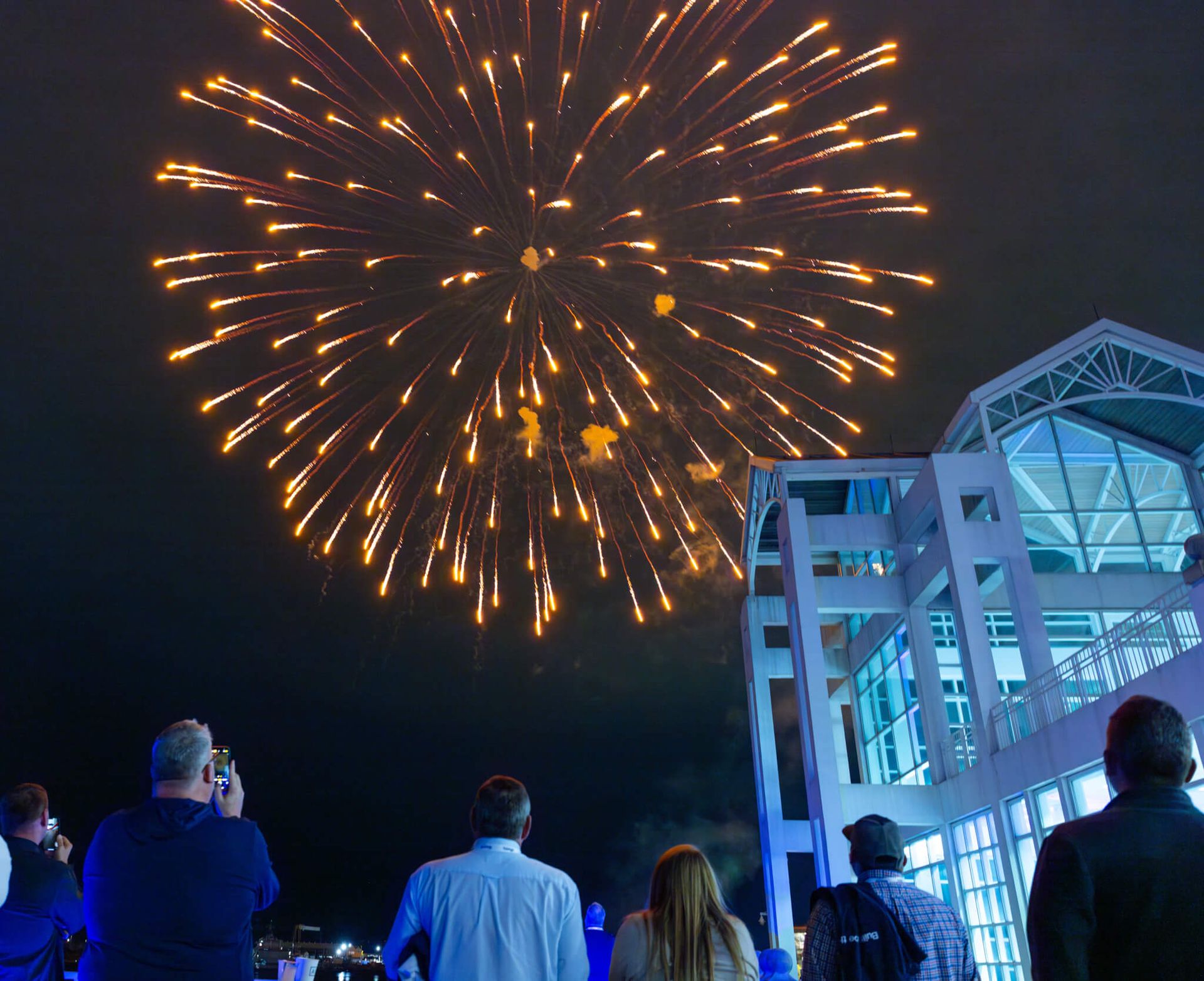 People watching golden fireworks above a lit modern building at night