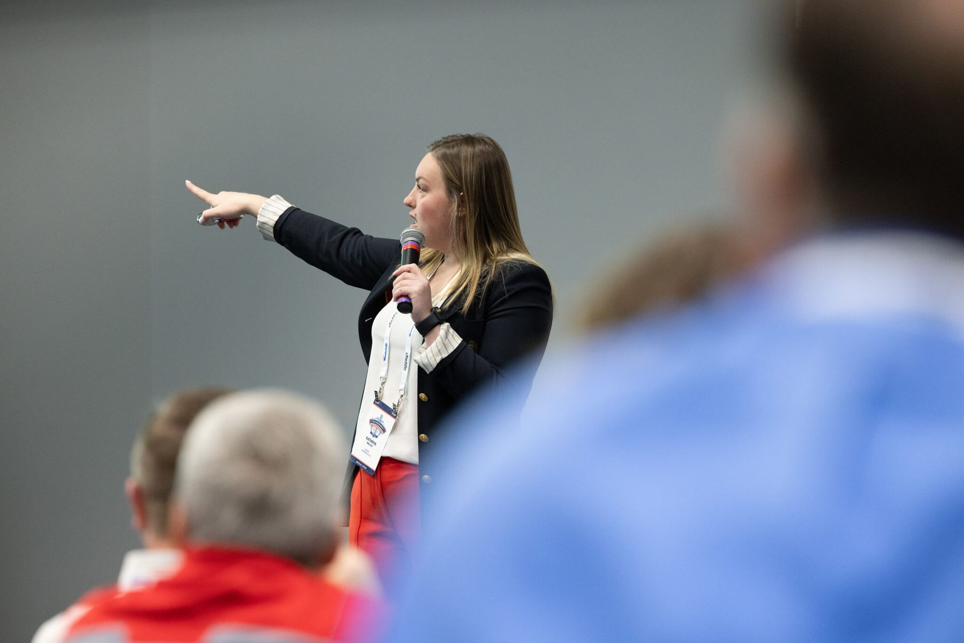 Speaker holding a microphone and pointing during a presentation, with audience members blurred in the foreground