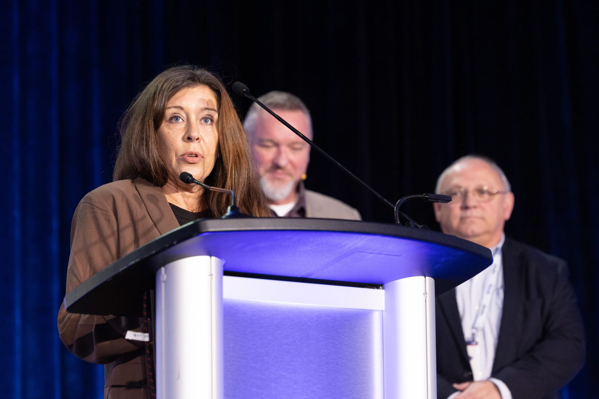 Woman speaking at a podium on stage, with two men standing behind her against a dark curtain.
