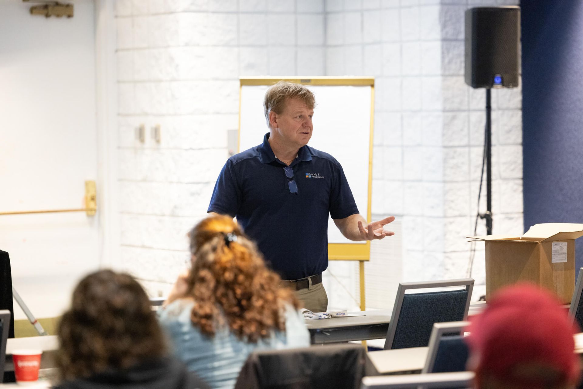 Speaker presenting in a classroom to seated attendees, with a laptop and projector screen nearby.