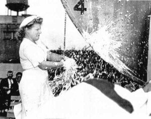 Worker spraying foam at a ship’s hull, with water splashing in a black-and-white photo.