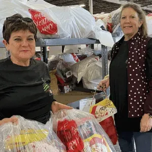 Two women stand by shelves of plastic bags in a storage area, one holding a large bag in front.