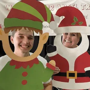 Two smiling people posing through elf and Santa cutout boards at a holiday event