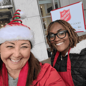 Two smiling volunteers in red coats and Santa hats pose outside a Salvation Army store.