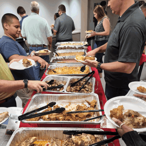 People serving food at a buffet table with trays of meat and sides at a crowded indoor gathering