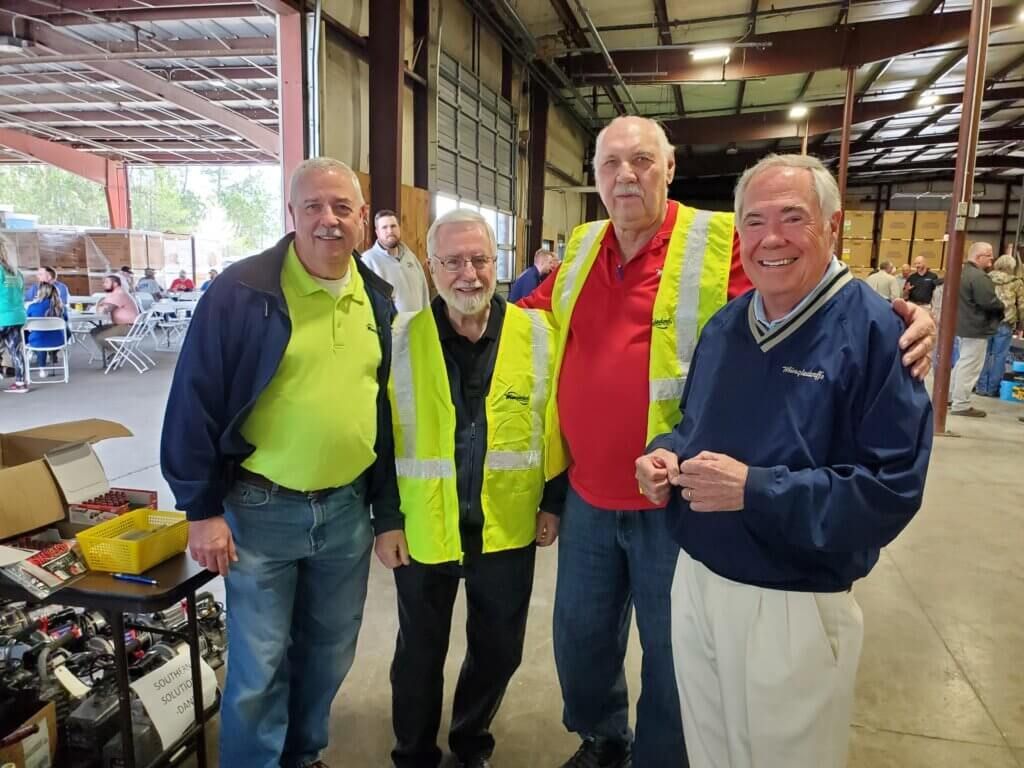 Three men smiling in neon safety vests inside a warehouse or workshop