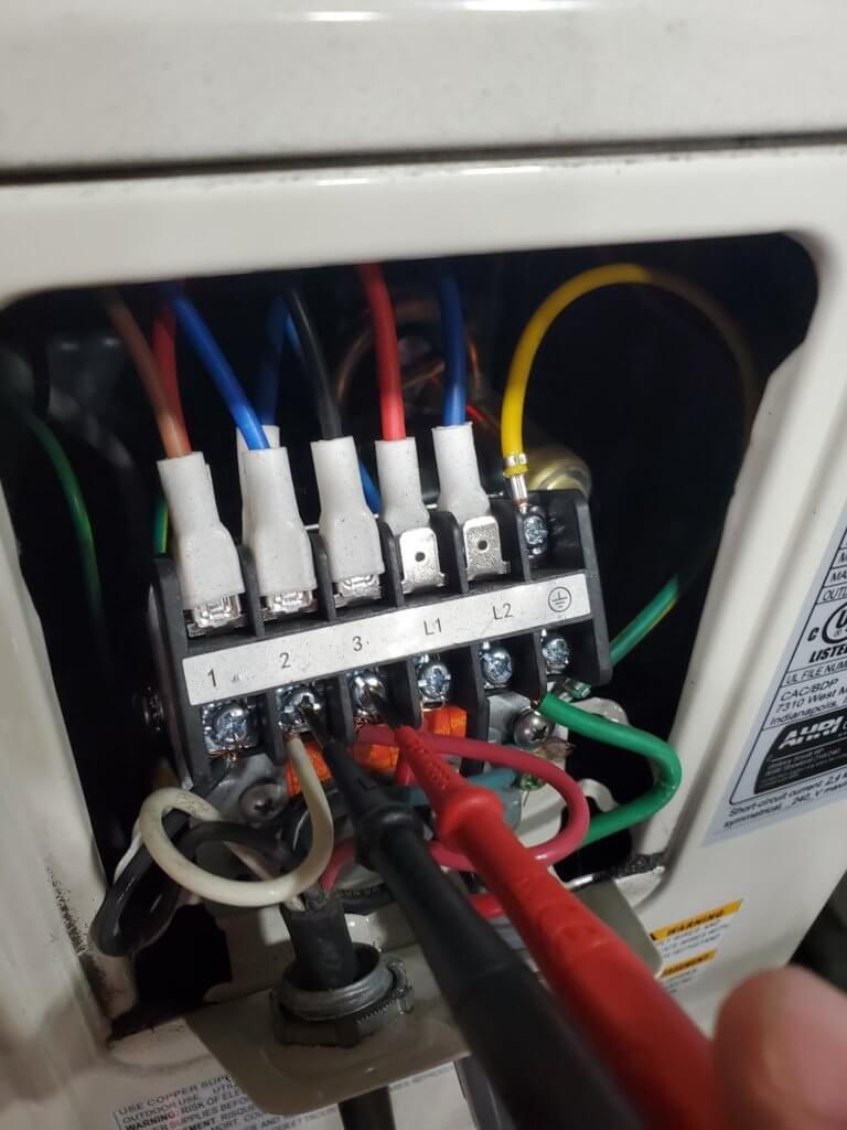 Electrical wiring terminal block inside an appliance, with colored wires and a hand holding red probes.