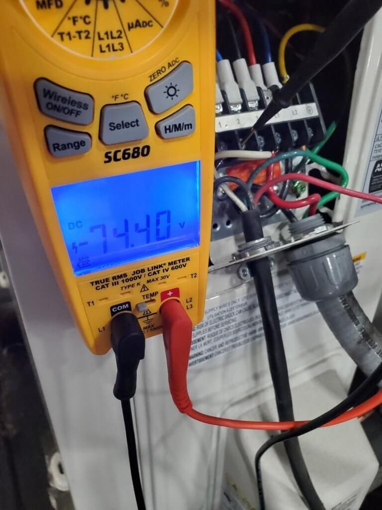 Yellow digital multimeter with red and black probes measuring wiring in an electrical panel; display shows 74.0V