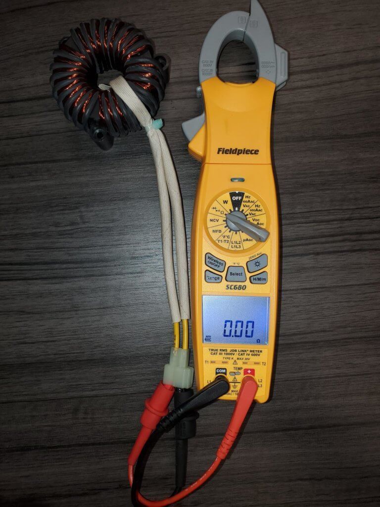 Yellow clamp meter with coiled current probe and red/black test leads on a dark surface, showing 0.0