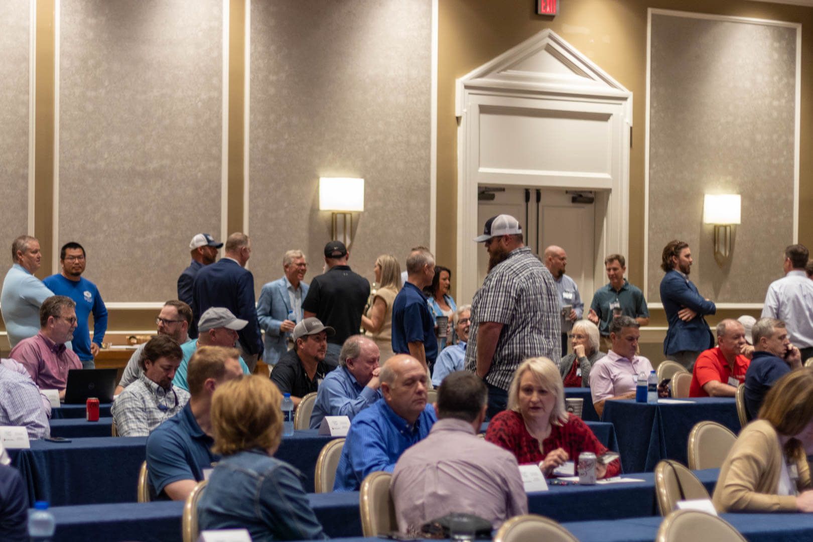 People seated and standing in a conference room with blue chairs and beige walls, gathered for an event