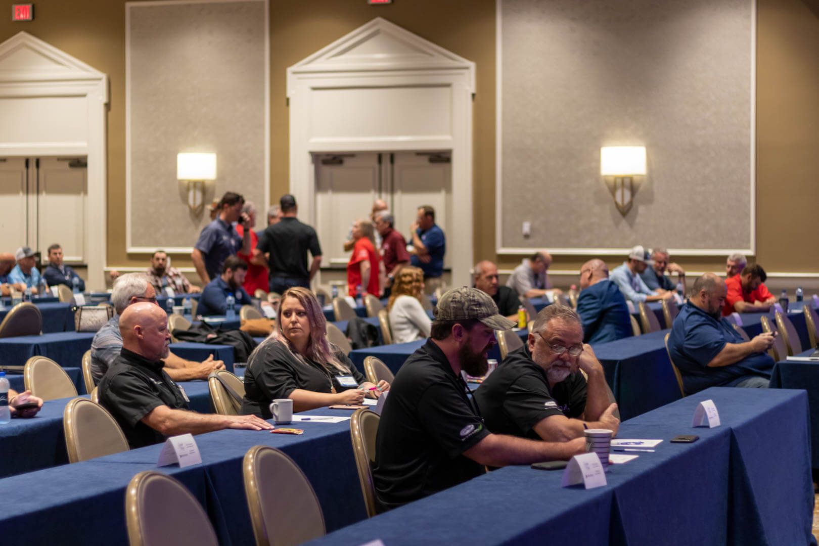 Conference attendees seated at blue-draped tables in a hotel ballroom, with people standing in the back.