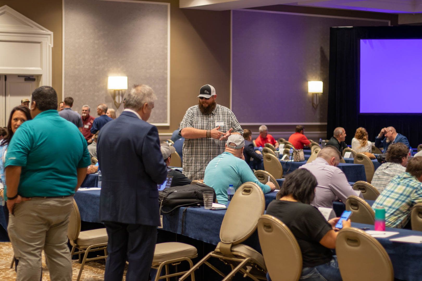 Conference attendees seated at tables in a hotel meeting room, with a presenter near a large screen.