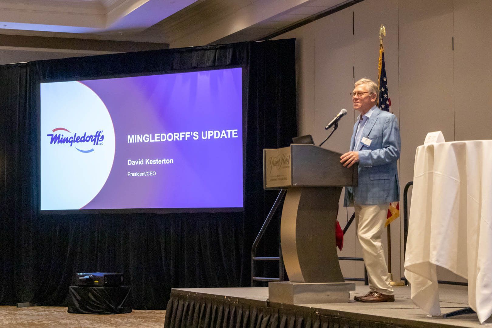 Speaker at podium beside purple presentation screen in a conference ballroom