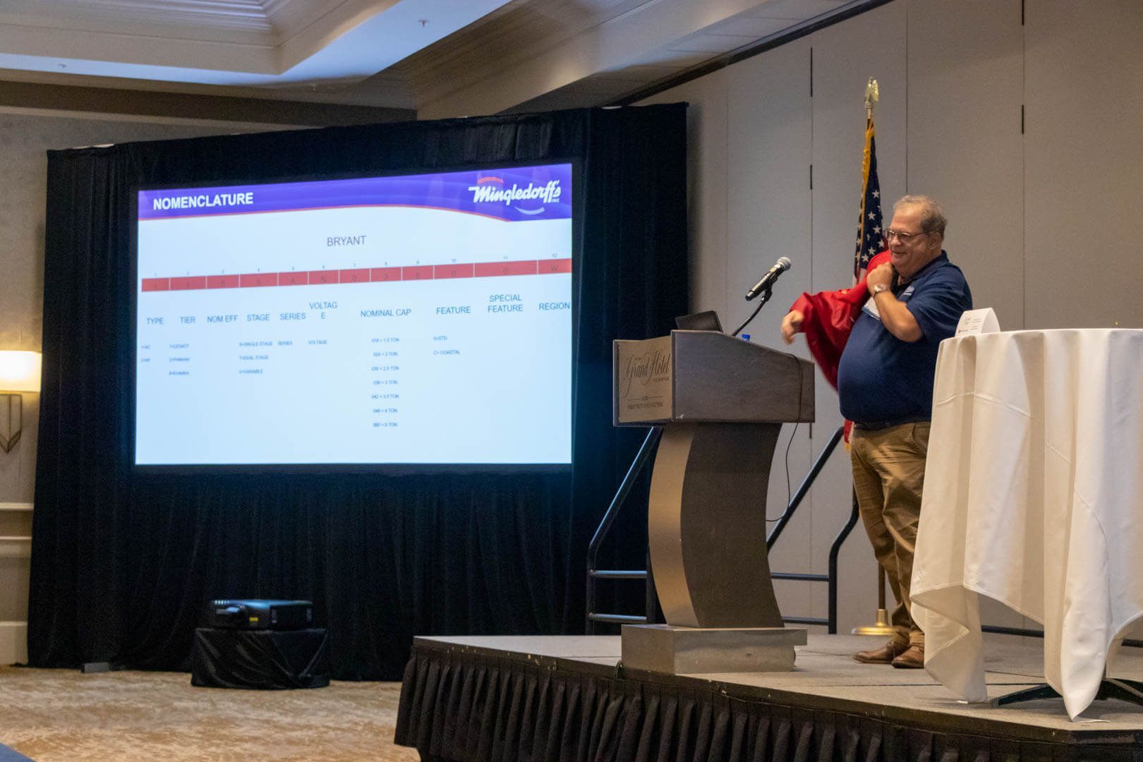 Man speaking at a podium beside a projected presentation screen in a conference room