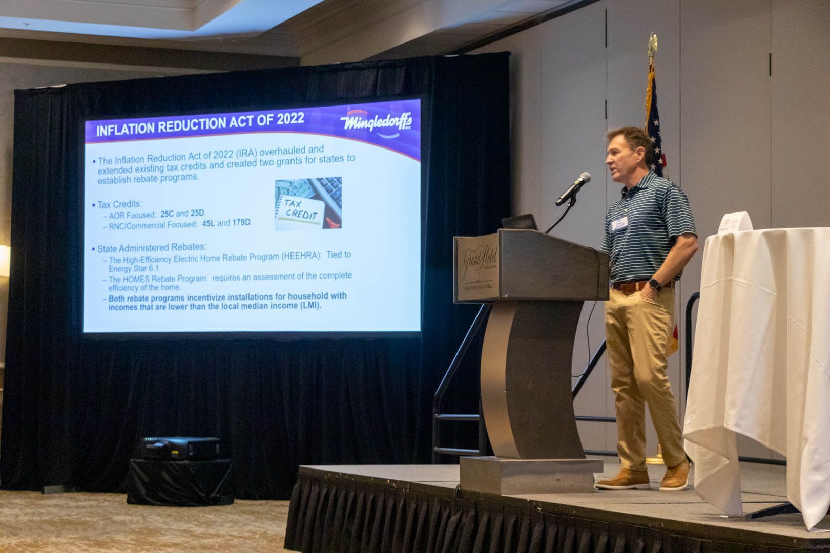 Speaker presenting at a conference beside a slide screen in a banquet hall