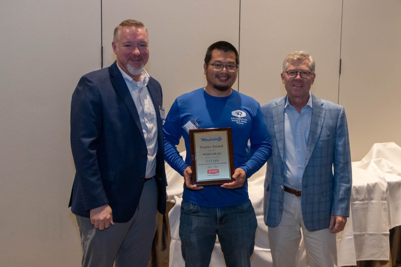Three men pose indoors; the center man holds a framed award plaque.