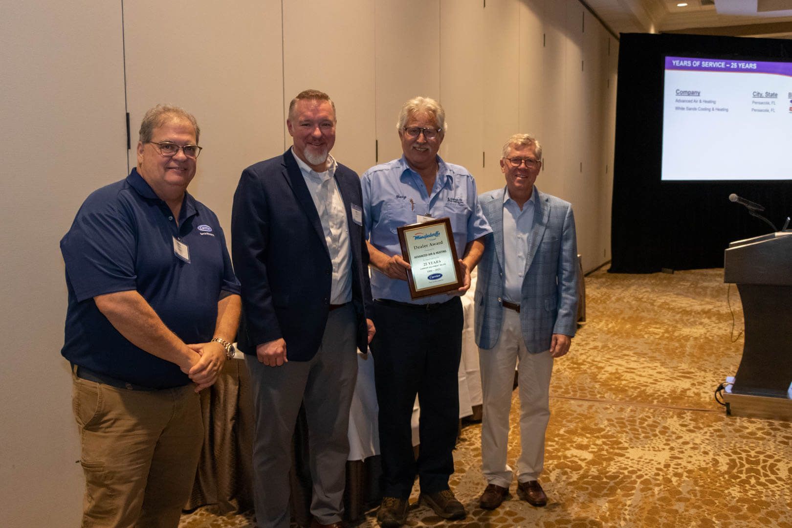 Four men at a conference hold an award plaque, posing beside a presentation screen.