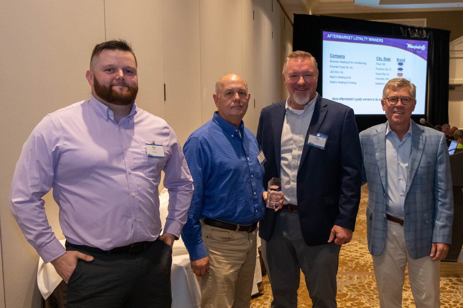 Four men posing indoors at a networking event, standing beside a presentation screen.