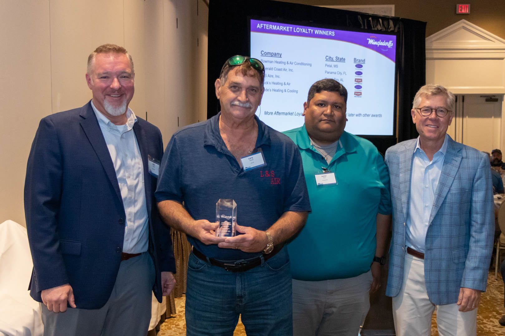 Four men posing at a conference, one holding an award plaque, with a presentation screen behind them.