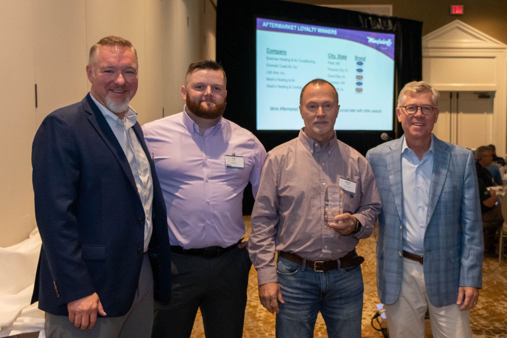 Four men posing at a conference event, with a presentation screen in the background.