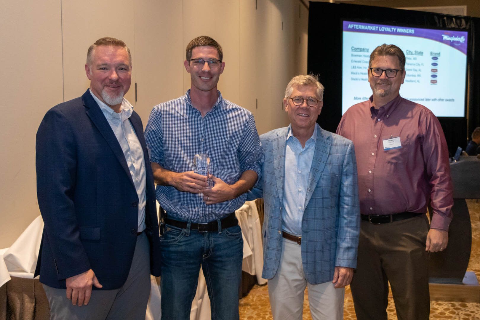 Four men posing together at an indoor event, smiling in front of a presentation screen.