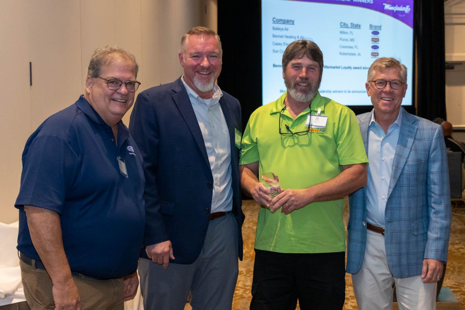 Four men standing together at a conference, smiling in front of a presentation screen.