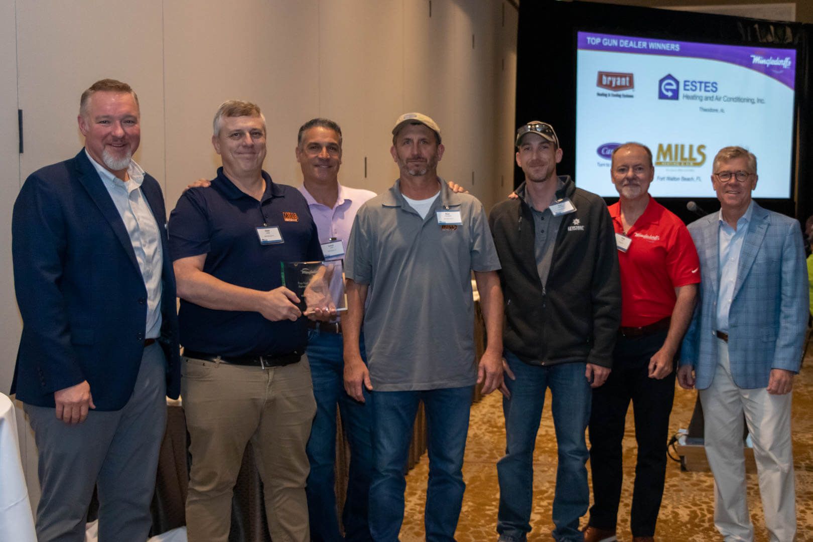Seven men posing at a conference event in front of a presentation screen and sponsor banner