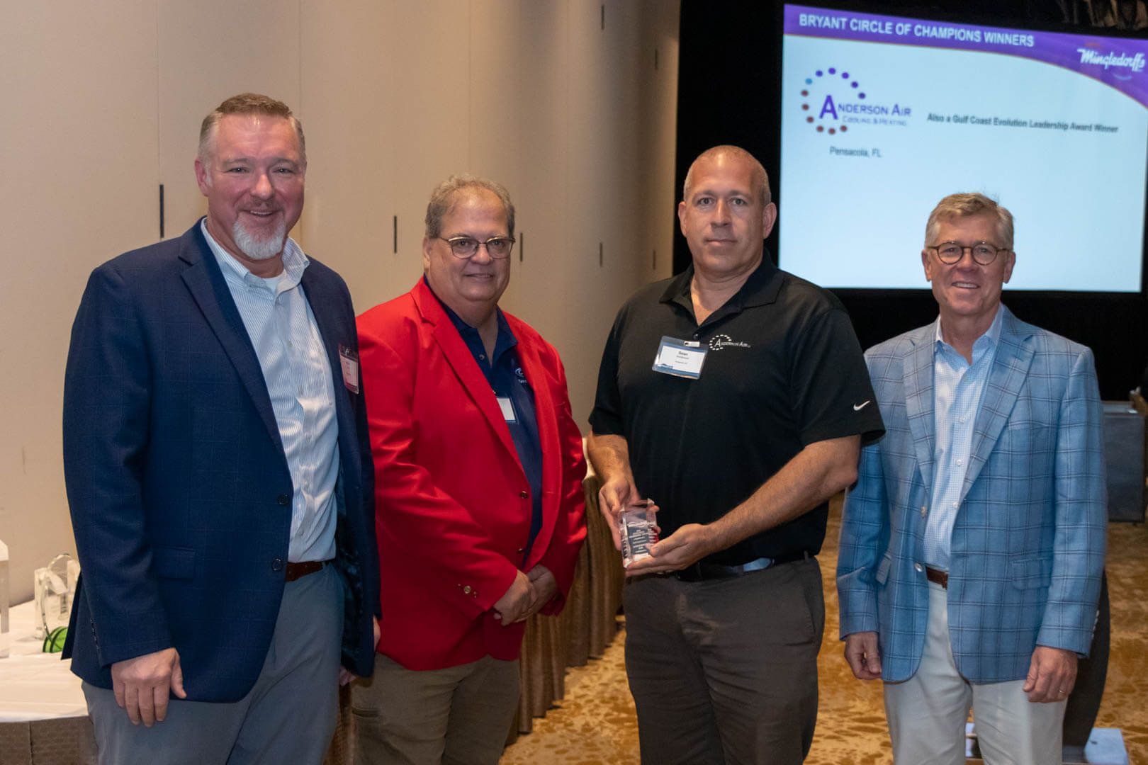 Four men posing at a conference, one holding an award plaque, with a purple event banner behind them.