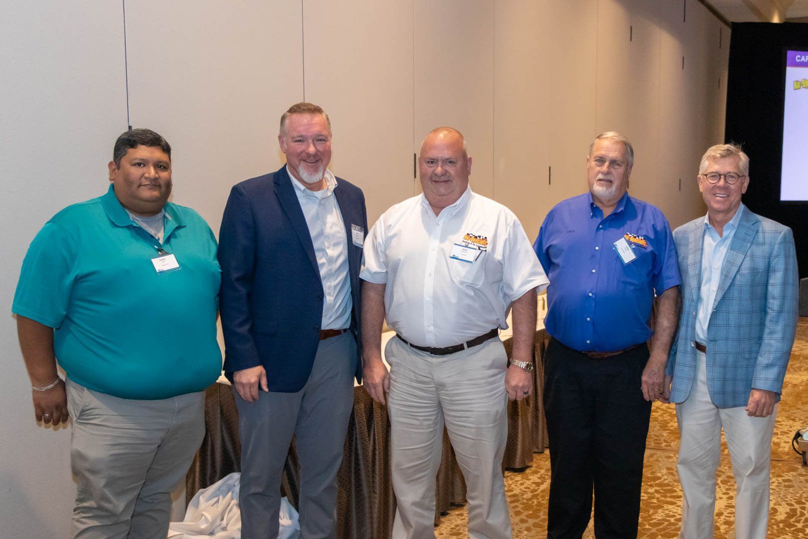 Five men stand together at an indoor event, dressed in business casual attire.