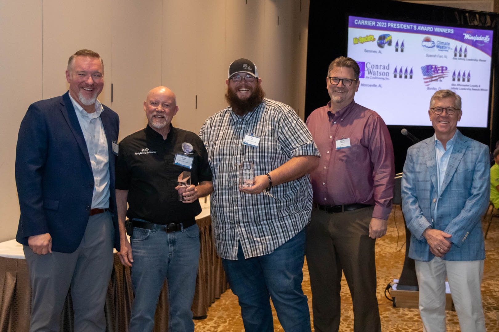 Five men posing at a conference, holding drinks, with a presentation screen in the background.