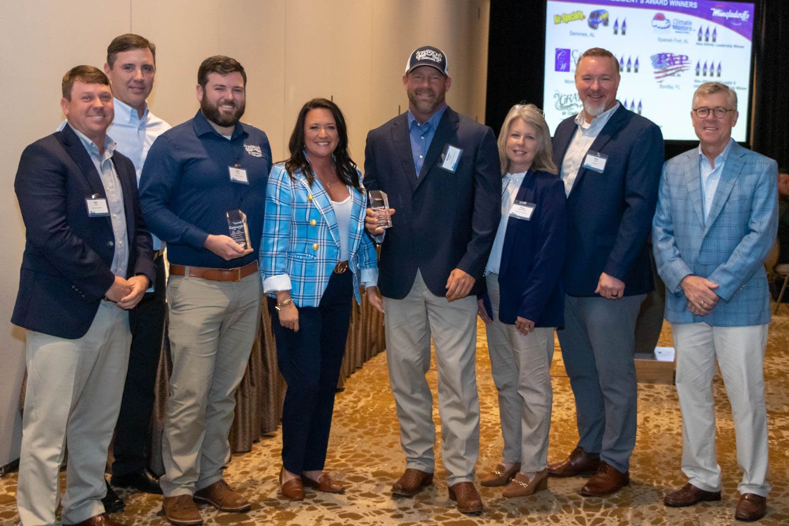 Seven people posing indoors at a conference, with one holding an award and a presentation screen behind them.