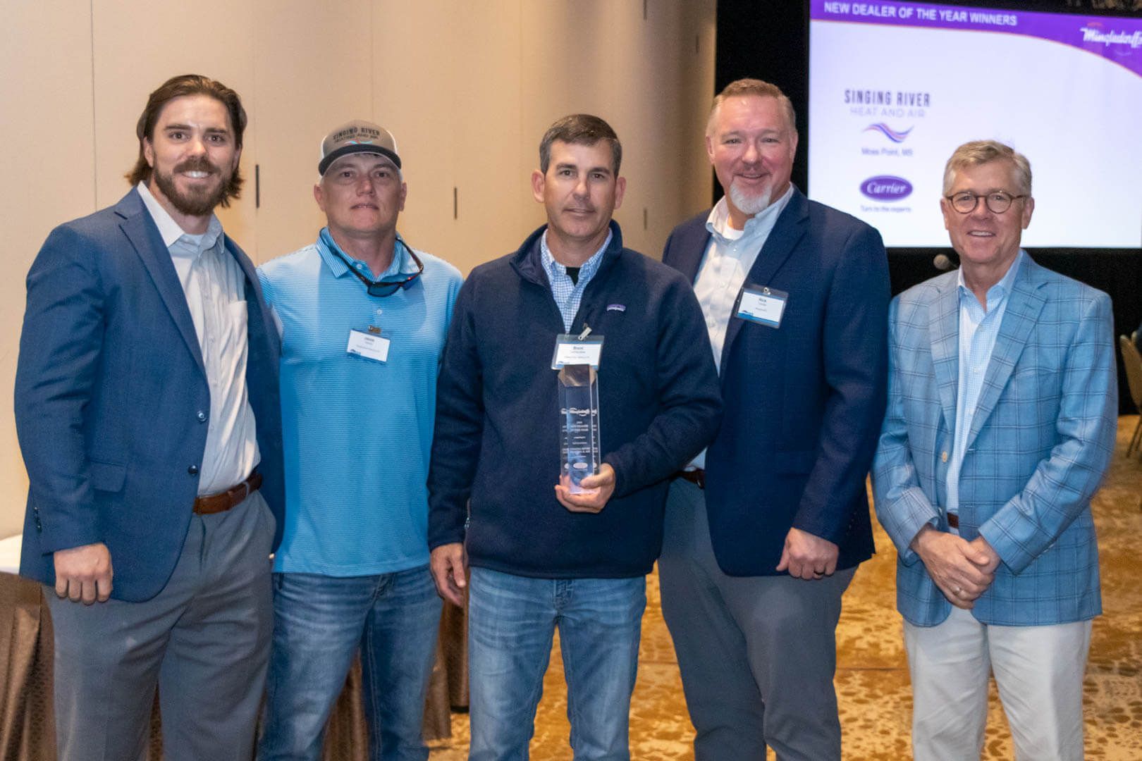 Five men pose on stage at an awards event, one holding a clear trophy plaque.