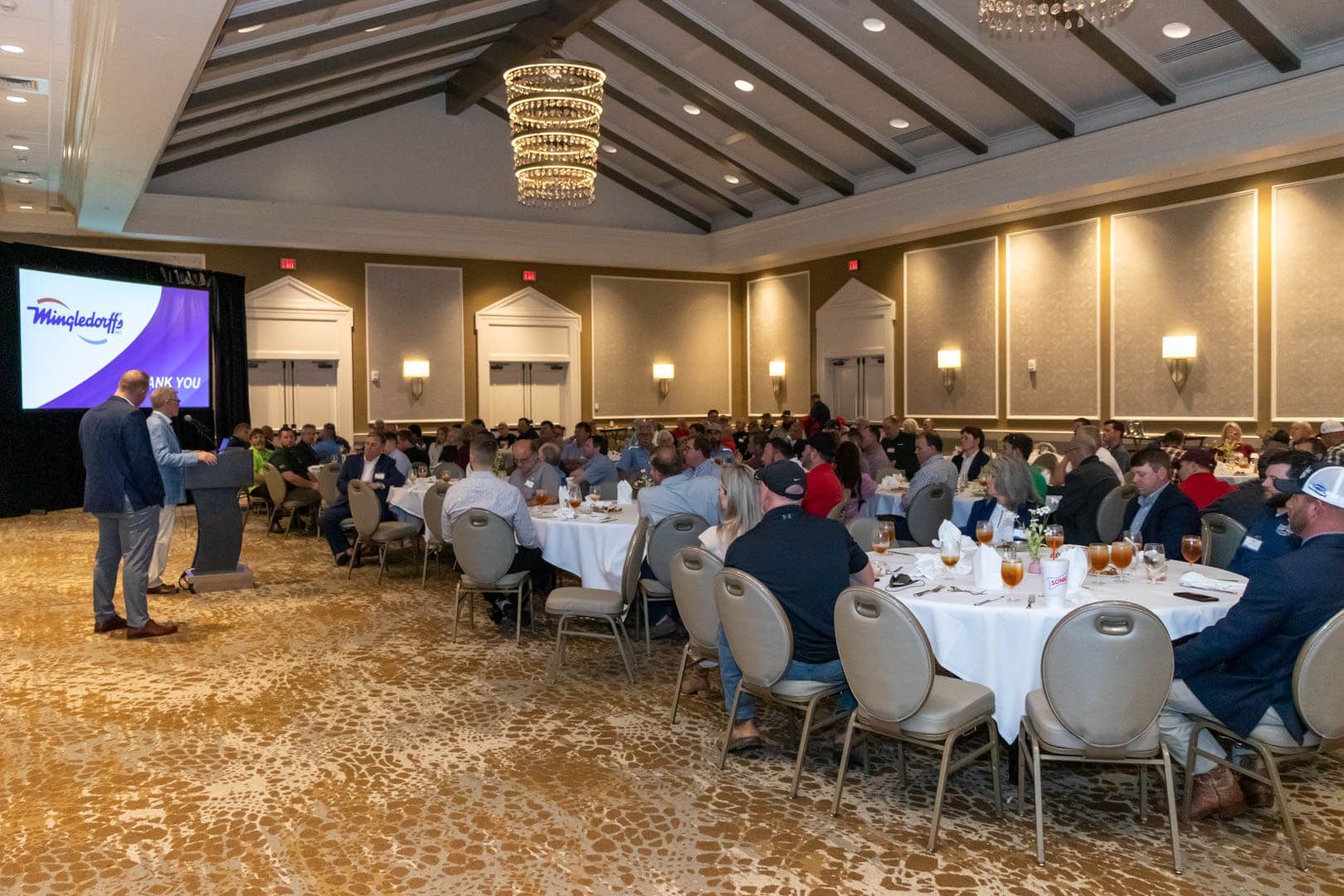 Conference banquet with seated audience facing a presenter and screen in a large hotel hall