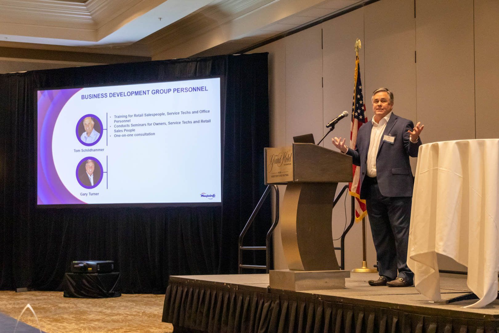 Presenter speaking on stage beside a projected slide at a conference, with a podium and U.S. flag.