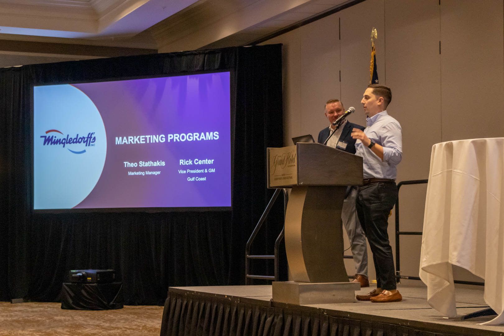 Presenter speaking at podium beside a projected “Marketing Programs” slide in a conference room