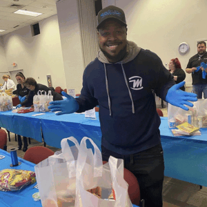 Smiling man in navy hoodie and blue gloves stands at a community table with gift bags and supplies
