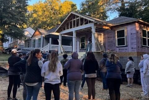 Group of people standing outside a small house with a porch and autumn trees in the background