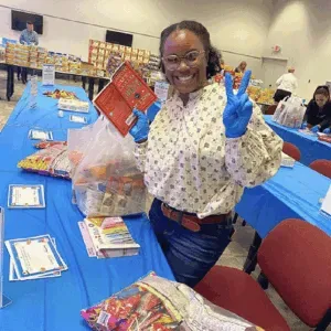 Person at a donation table in a blue-carpeted room, smiling and holding up packages with gift bags nearby