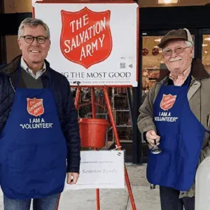 Two volunteers in blue aprons stand beside a Salvation Army donation kettle.