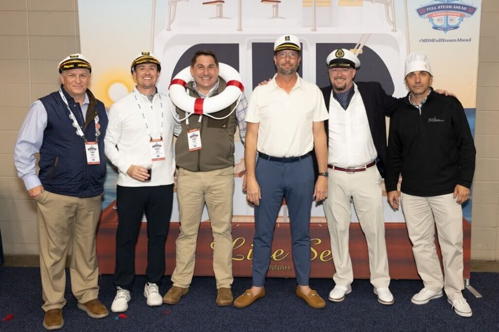 Six men posing indoors in front of a large white aircraft nose display.