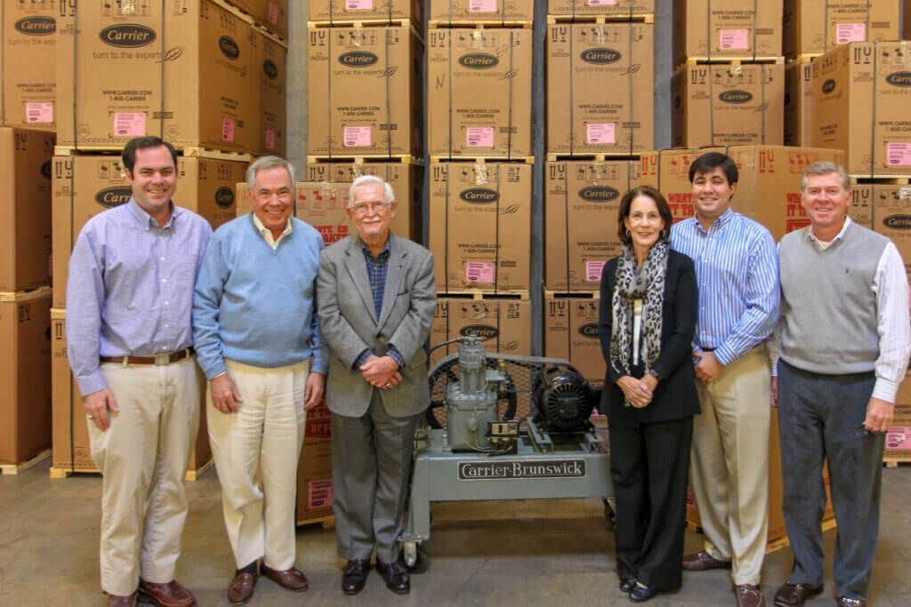Six people standing in front of stacked cardboard boxes in a warehouse, with a machine in the center.