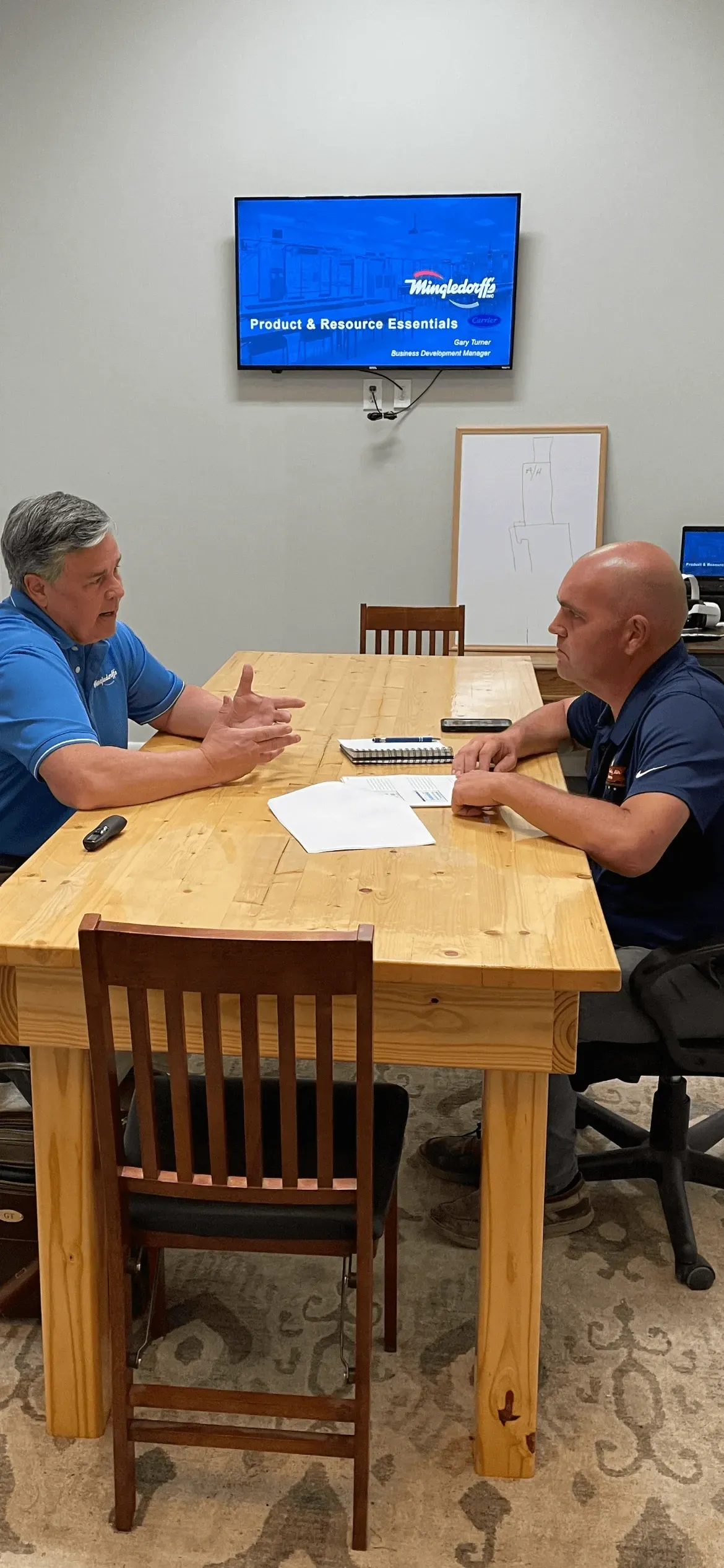 Two people sit at a wooden table in a room, talking beneath a wall-mounted TV with a blue screen.