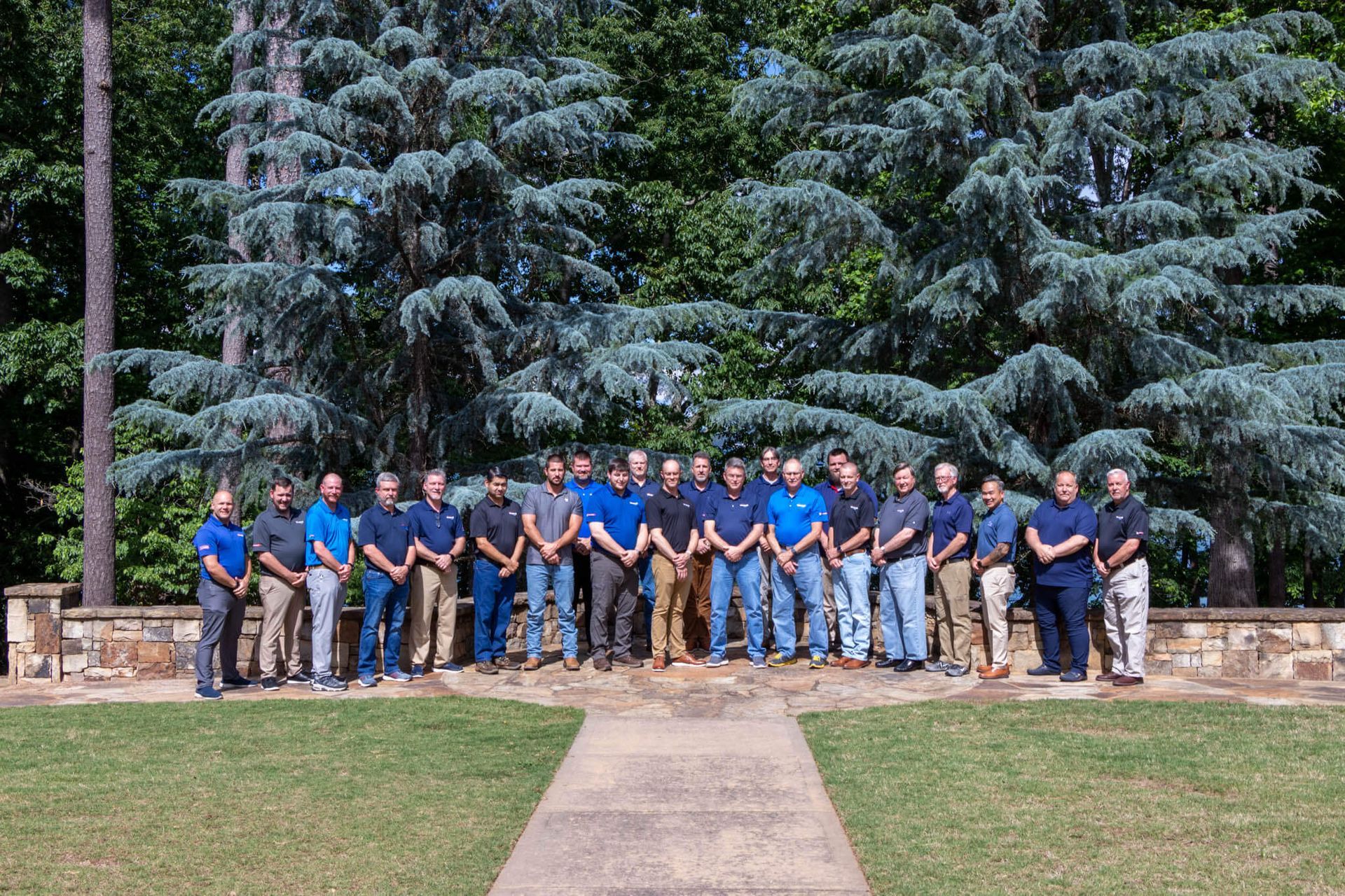 Group of people in blue shirts and jeans posing outdoors in front of tall evergreen trees