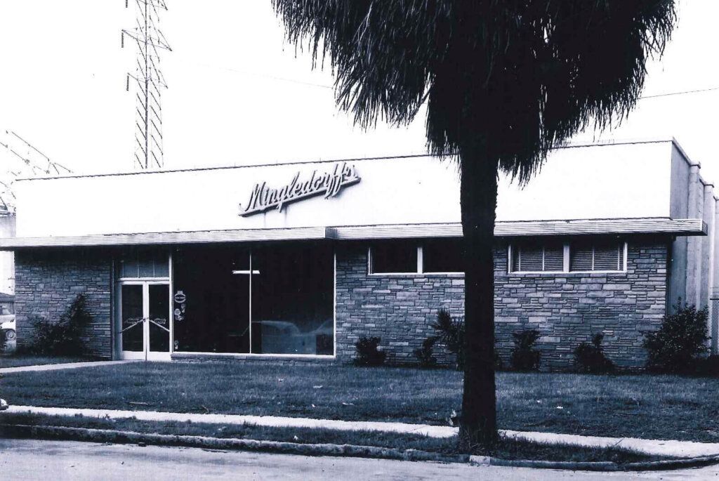Small storefront with stone facade and a sign, partly obscured by a tree in front.