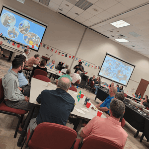Conference room meeting with seated attendees, flags on tables, and projected slides at the front.