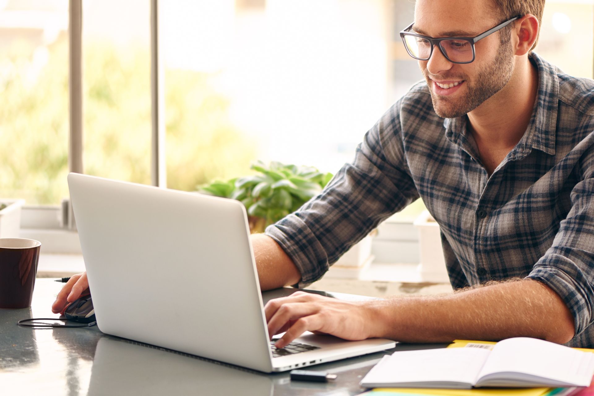 Smiling man in glasses working on a laptop at a desk with papers and a coffee mug